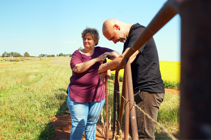 Two people leaning against a rusted gate in a field with canola flowers in the background.