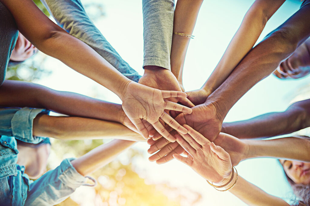 Several hands reaching and touching each other in the center of the frame with a warm light glow in the background.