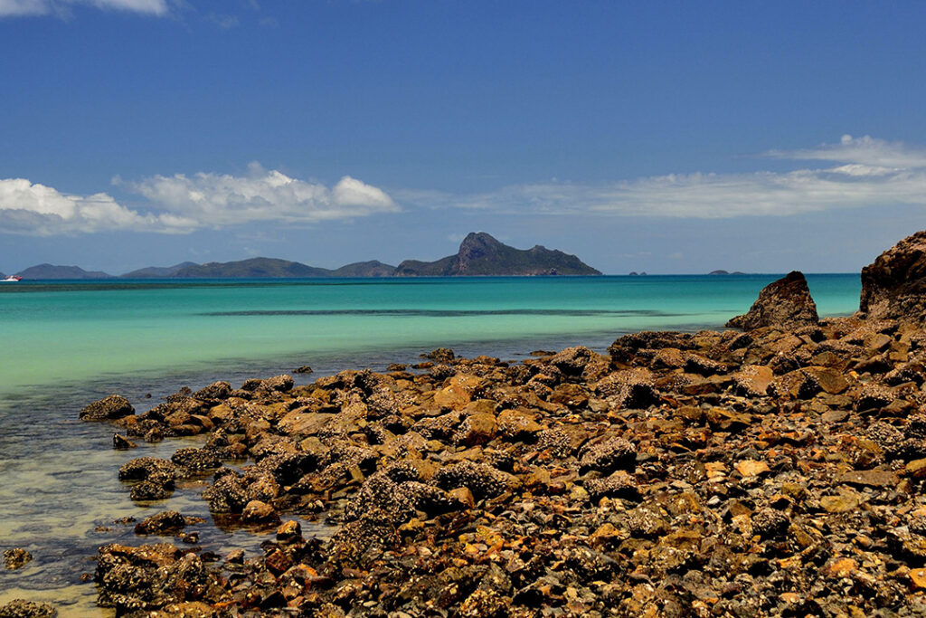 Looking across a rocky beach to turquoise water with a mountain island in the background.