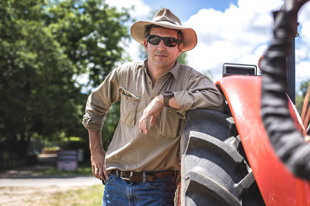 A person wearing a wide brim hat leaning against a tractor wheel in the country.